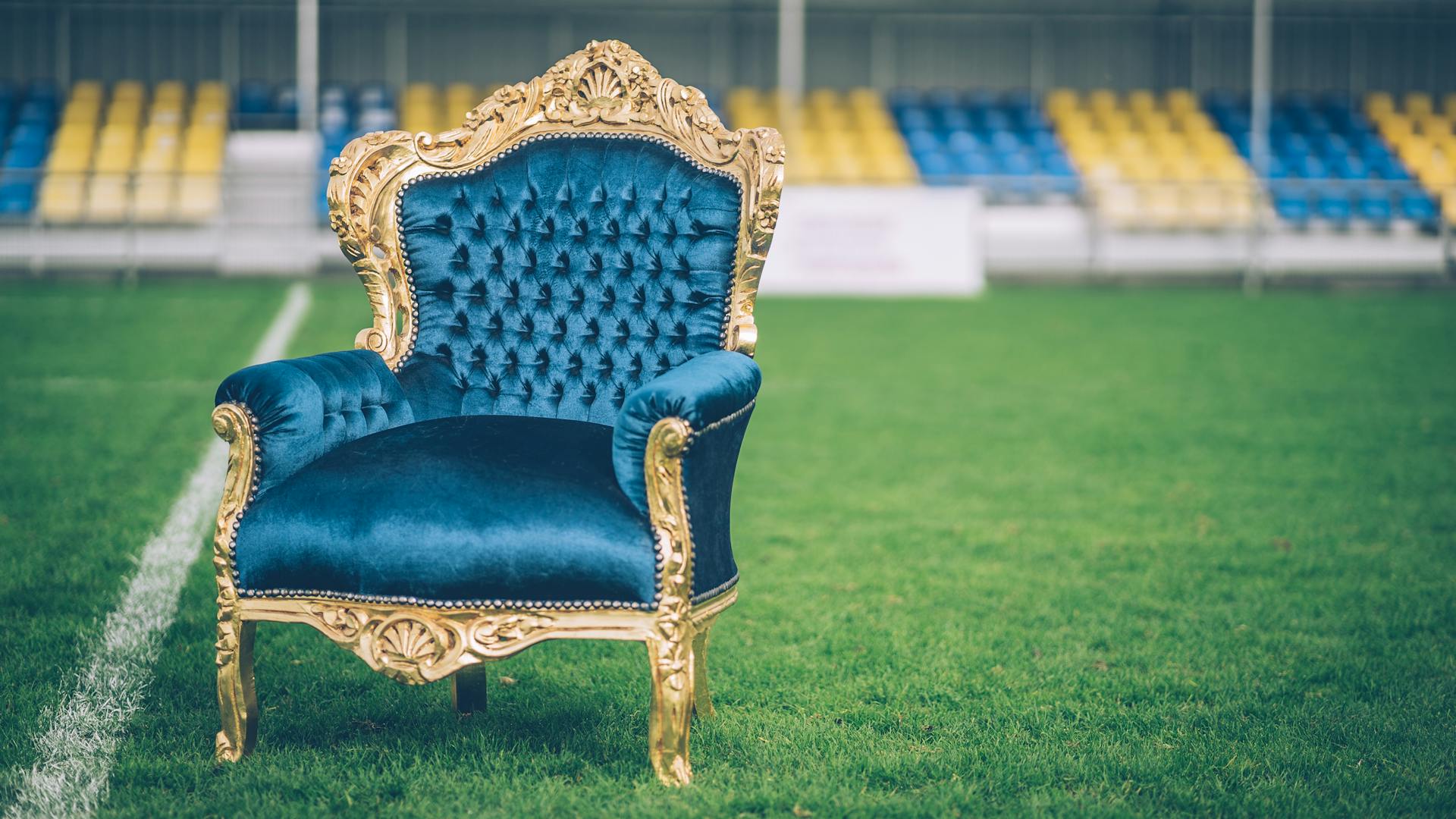 A luxurious blue velvet and gold armchair sits prominently on a sports field.