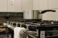 Saucepan on rack of old gas stove with valves in row and sticker with attention symbol against fabric