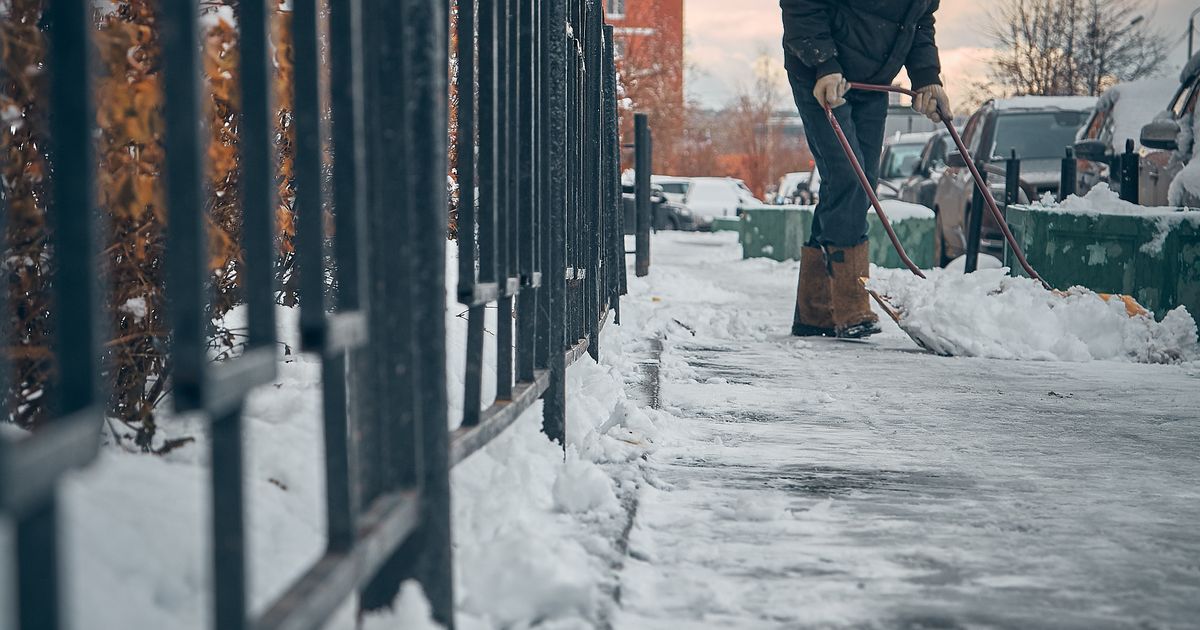 Mann in Winterkleidung schaufelt Schnee vom Gehweg entlang eines Metallzauns, Symbolbild für Gehwegräumung