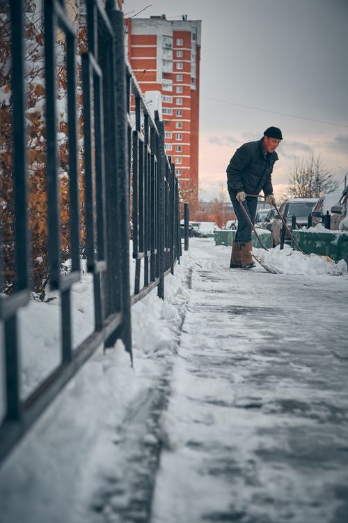 Free A man wearing winter clothing shovels snow from a sidewalk by a metal fence in front of a building. Stock Photo