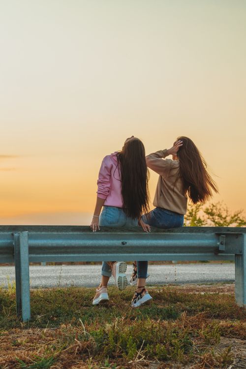 Free Back view of anonymous female friends with long hair in casual clothes sitting together near roadway Stock Photo