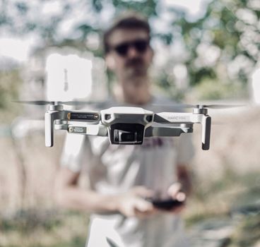 Drone in focus hovering outdoors with operator blurred in the background.