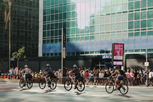 Free Cyclists ride past a crowded building entrance in a sunny urban setting, showcasing city life and mobility. Stock Photo