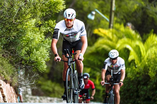 Full body of faceless men in helmets and eyeglasses riding bikes on road surrounded by verdant trees and bushes in sunlight on blurred background