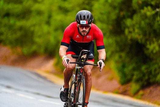 Full body of tanned young slim bicyclist in tracksuit and helmet riding bike along tall green trees in daytime on blurred background