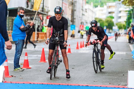 Two cyclists in a street race, wearing helmets and sportswear, surrounded by cones.