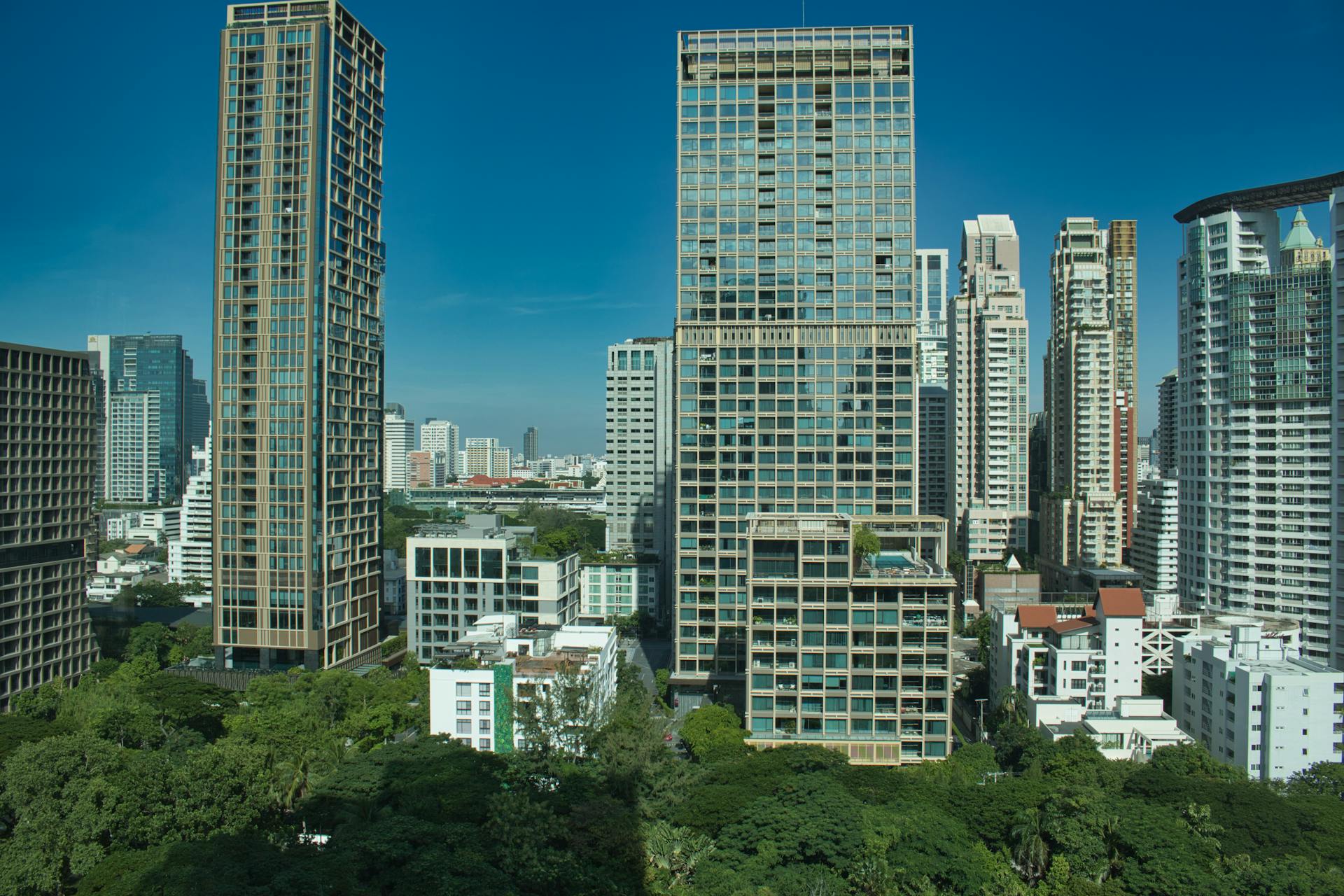 Explore a vibrant Bangkok cityscape with modern high-rise buildings and lush greenery under a clear blue sky.