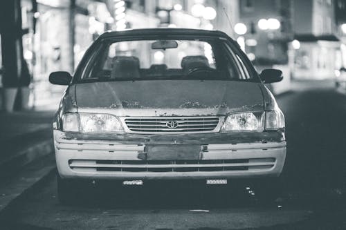 Black and white of old timer automobile with headlights and rusty hood parked on asphalt road with blurred lights in city on background