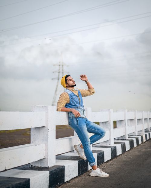 Free Trendy man in denim overalls and yellow beanie posing stylishly outdoors against a bridge railing. Stock Photo