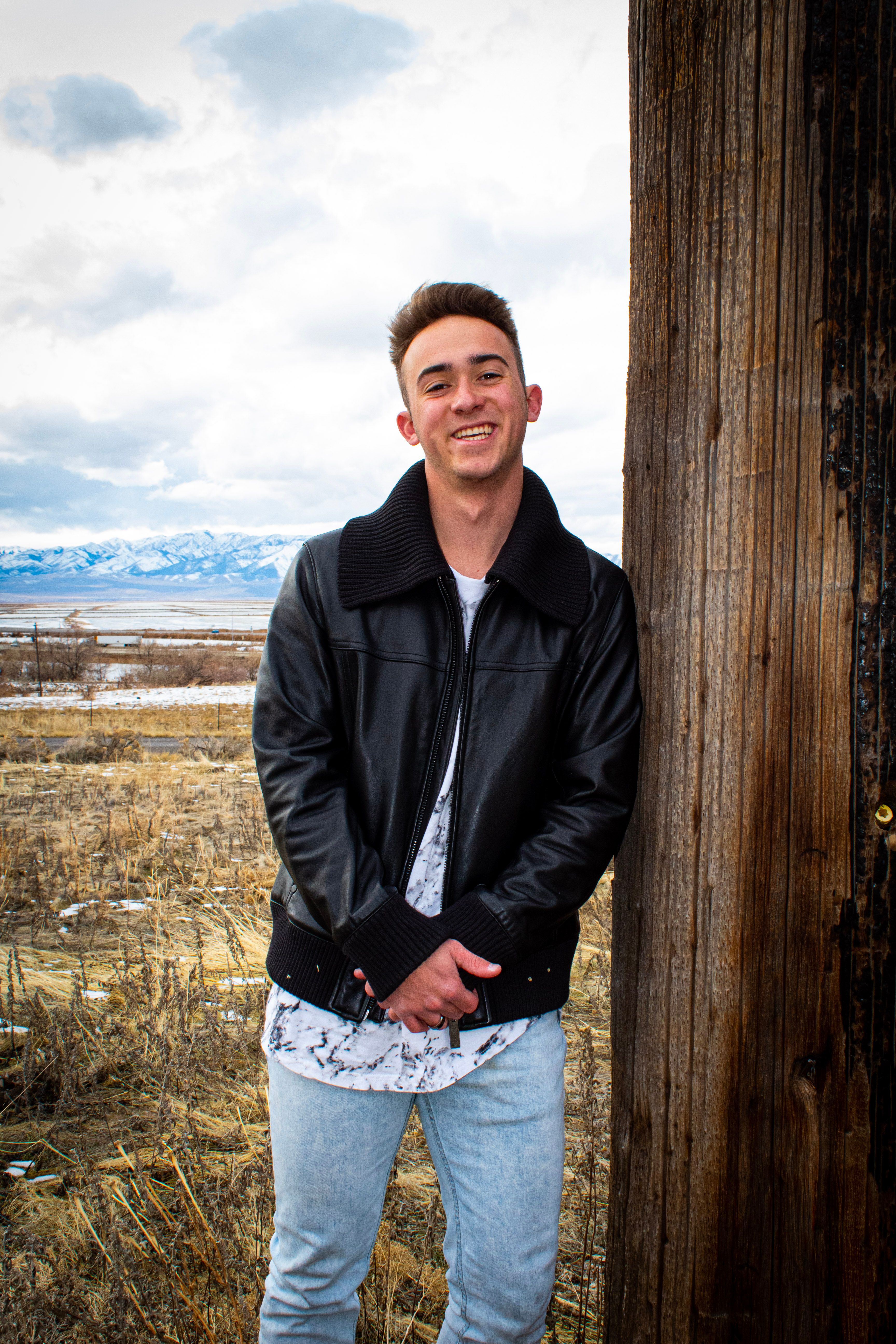 Smiling young man in leather jacket posing outdoors against rustic landscape.