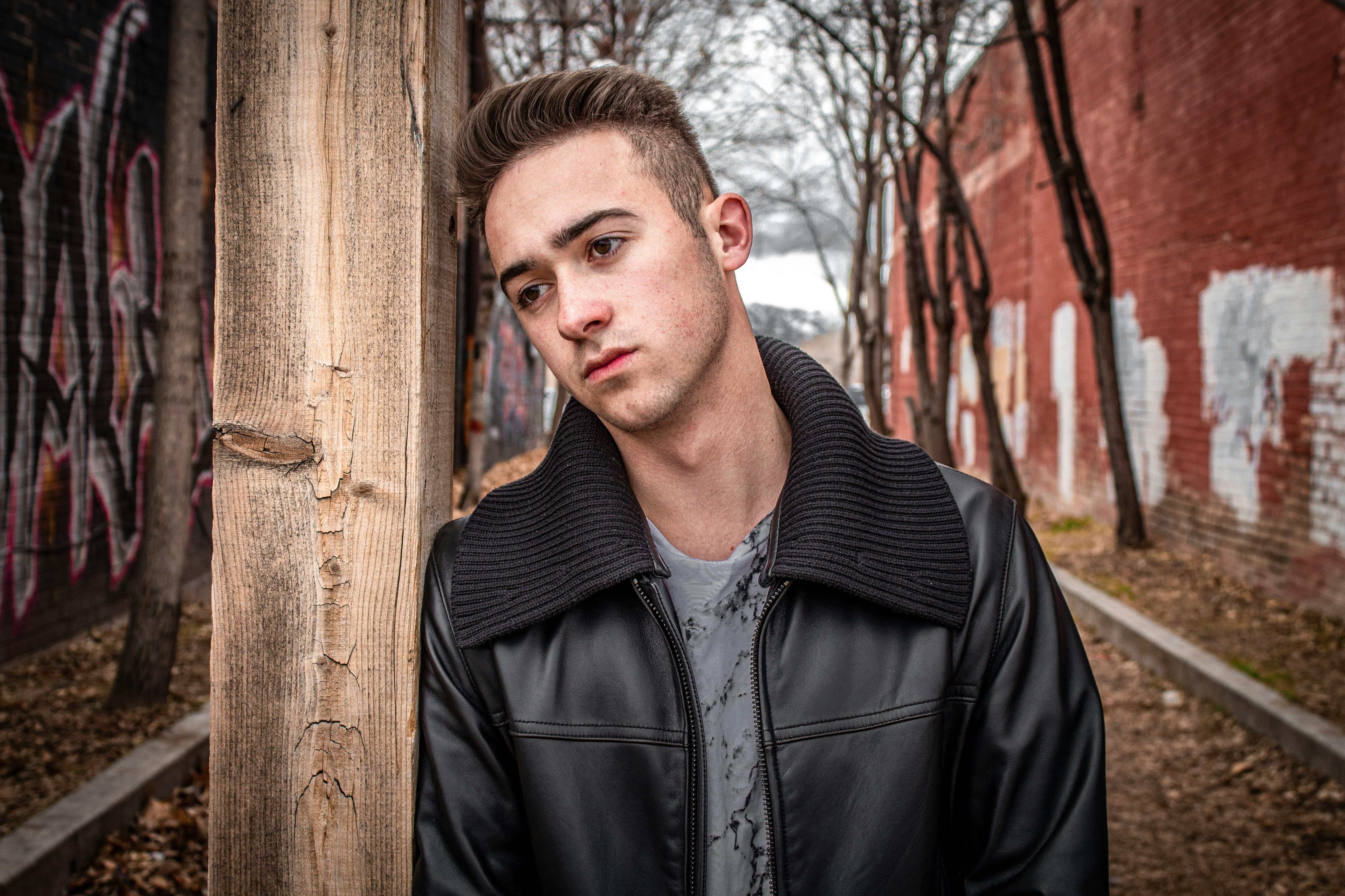Portrait of a young man in a black jacket leaning against a wooden post with graffiti background.