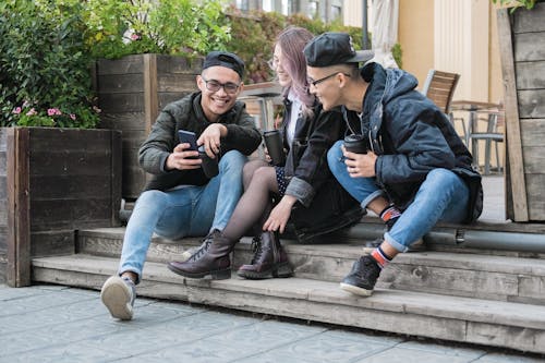 Cheerful ethnic friends browsing smartphone and sitting on cottage stairs