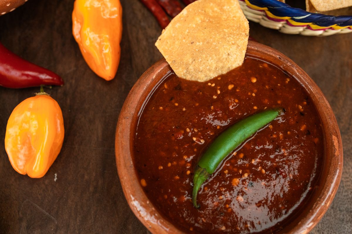 Clay bowl of dark red roasted chile salsa with a whole green serrano and tortilla chip
