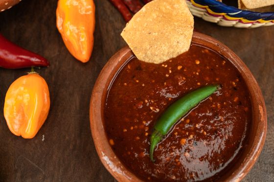 Clay bowl of dark red roasted chile salsa with a whole green serrano and tortilla chip