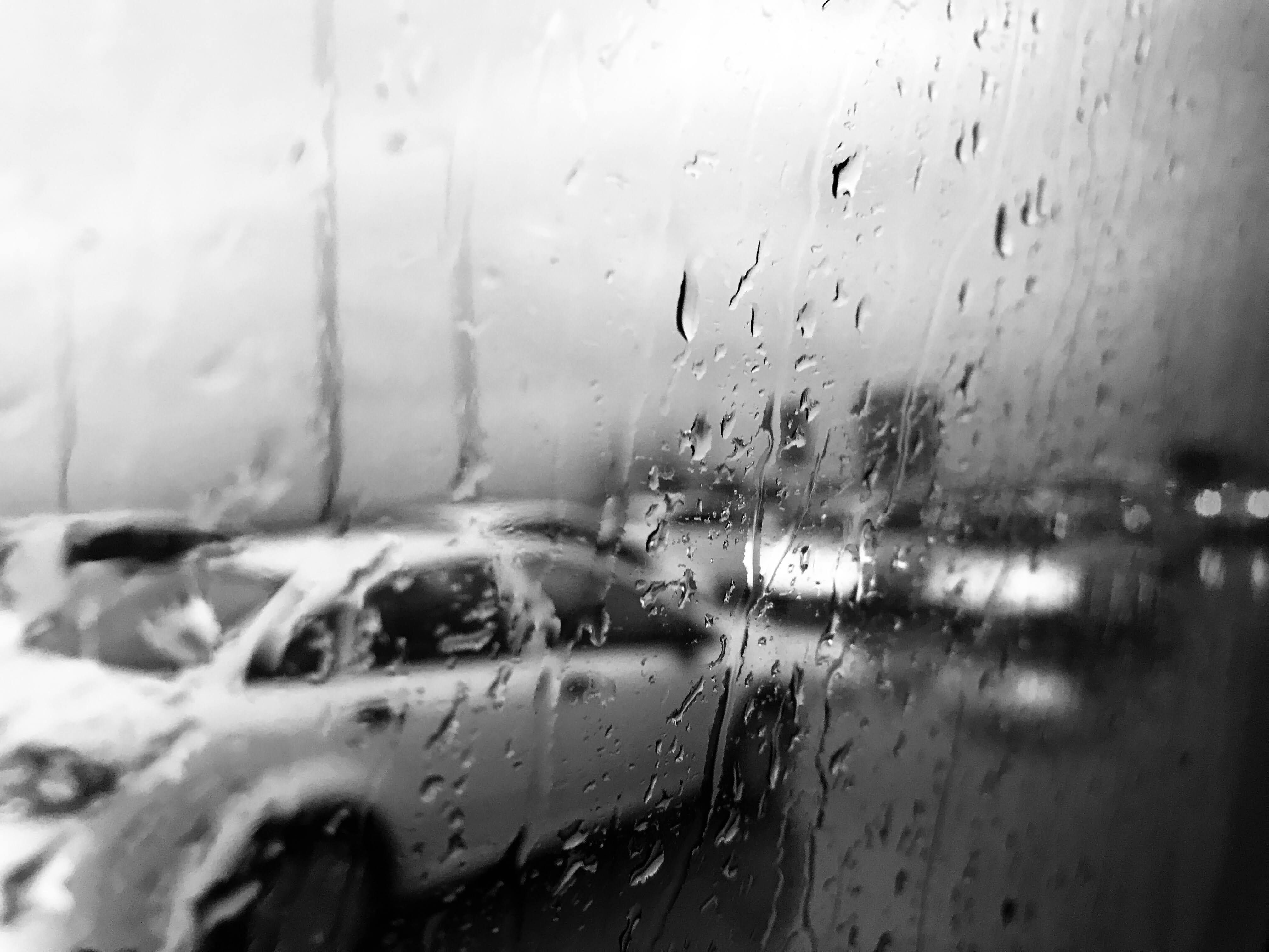 A black and white image capturing a car seen through rain-speckled glass.