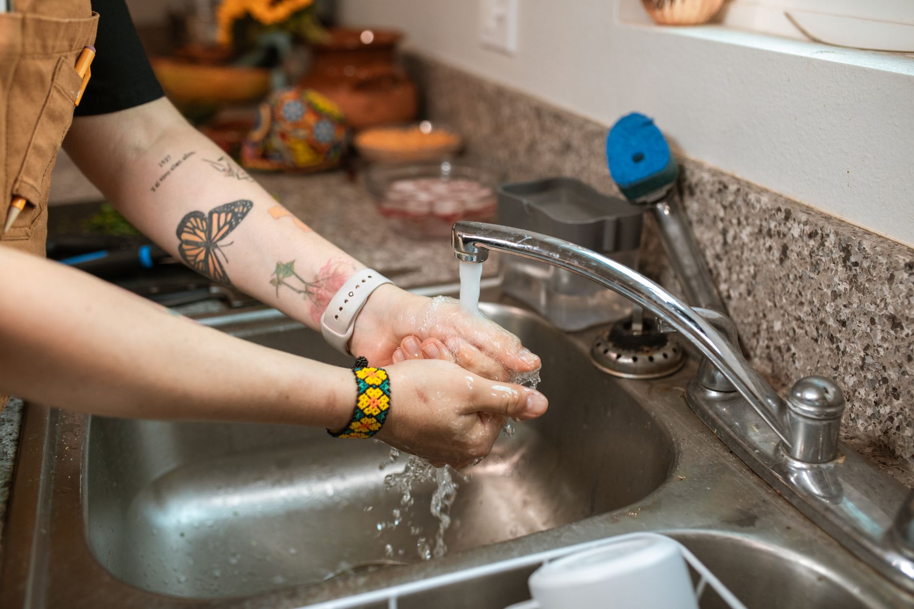 Charlotin Plumbing Services - Close-up of tattooed hands being washed under a kitchen faucet, emphasizing cleanliness and hygiene.