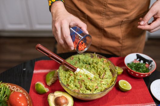 High angle crop unrecognizable chef wearing brown apron adding cut tomatoes into bowl with delicious savory guacamole placed on table