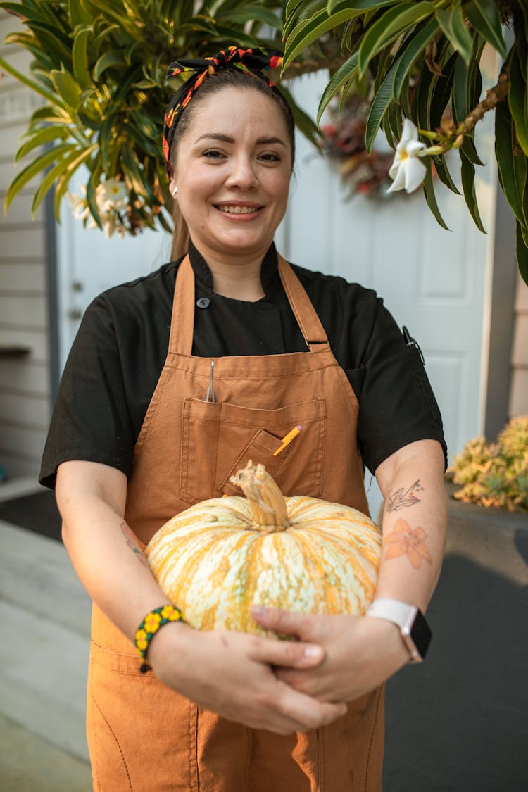 A Woman In Brown Apron Smiling While Holding A Pumpkin