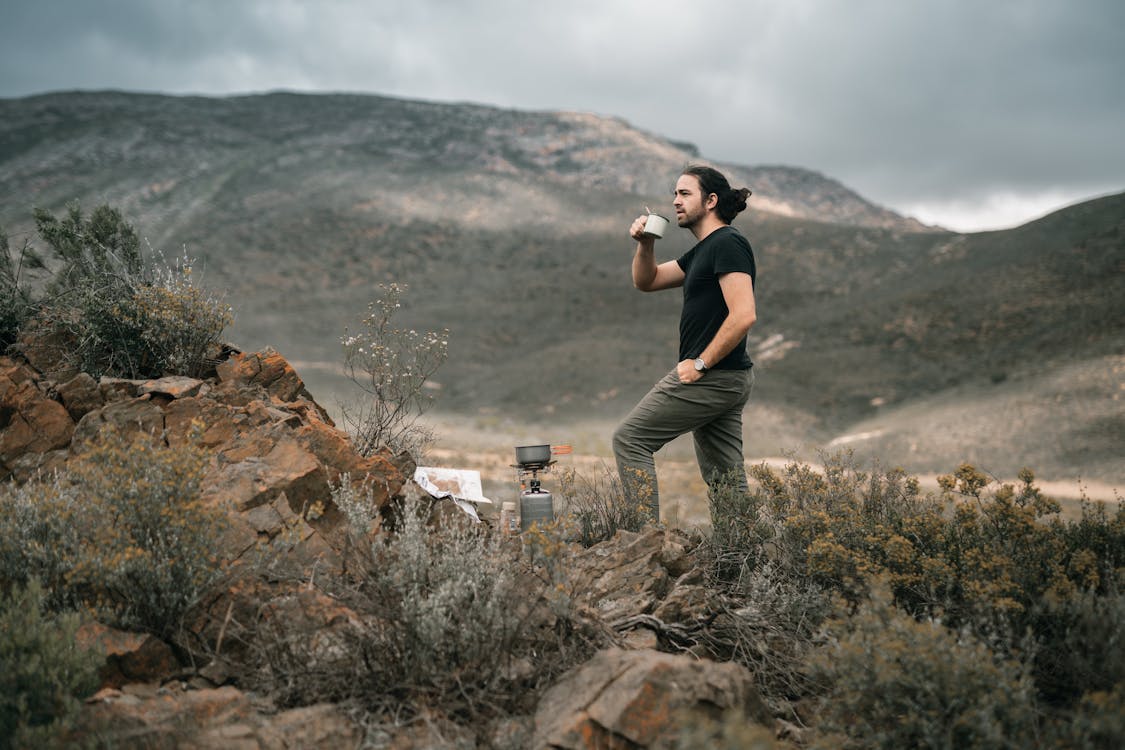 A Man in Black Shirt Standing while Drinking Coffee Near the Mountain
