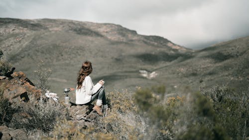 Free A Woman in White Sweater Sitting on the Rock while Looking at the Mountain Stock Photo
