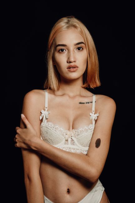 Young gentle woman in lace bra and white panties touching arm while looking at camera on black background