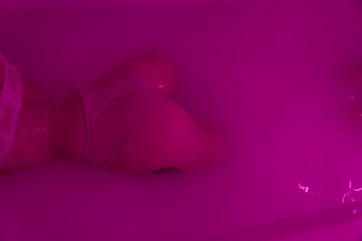 From above back view of crop anonymous seductive female in bra and thong lying in water in neon light