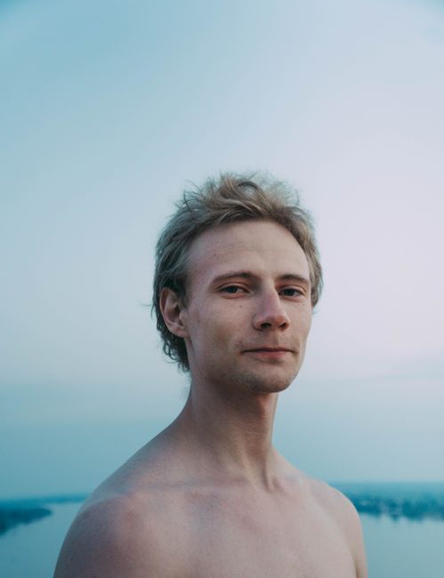 Young friendly male with naked torso and uncombed hair looking at camera against river in daytime