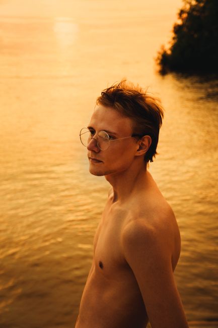 Side view of young male with naked torso in eyewear looking away against rippled lake in evening