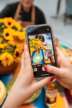 Close-up of hands using smartphone to photograph a summer table with bright sunflowers and food.