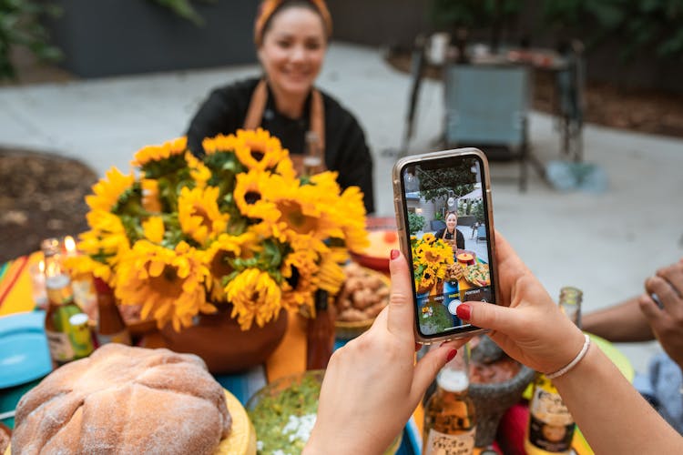 A Person Holding A Mobile Phone While Taking Picture Of A Smiling Woman Near The Flowers