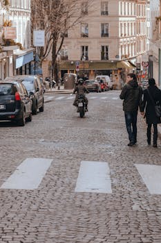 A lively urban street scene with people walking and a motorcyclist riding down a cobblestone road.