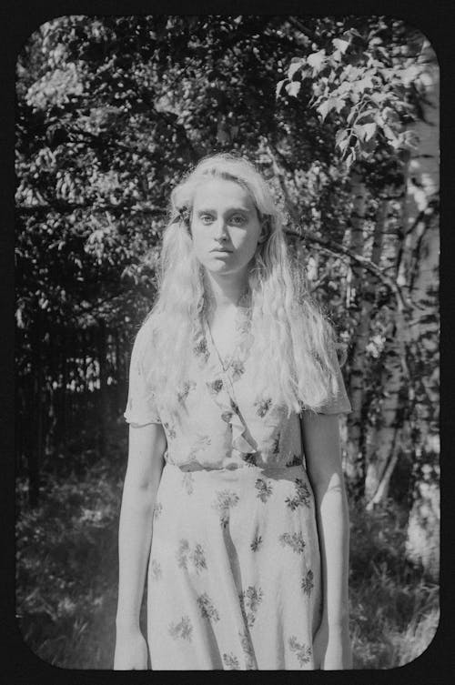 Black and white of young tender female in ornamental outfit looking at camera against birches in sunlight