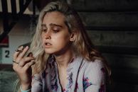 一位金发女子身穿花卉上衣，手持香烟，在楼梯上陷入沉思
