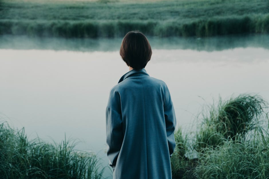 Back view of anonymous person in coat admiring river reflecting lush grass in summer in daylight