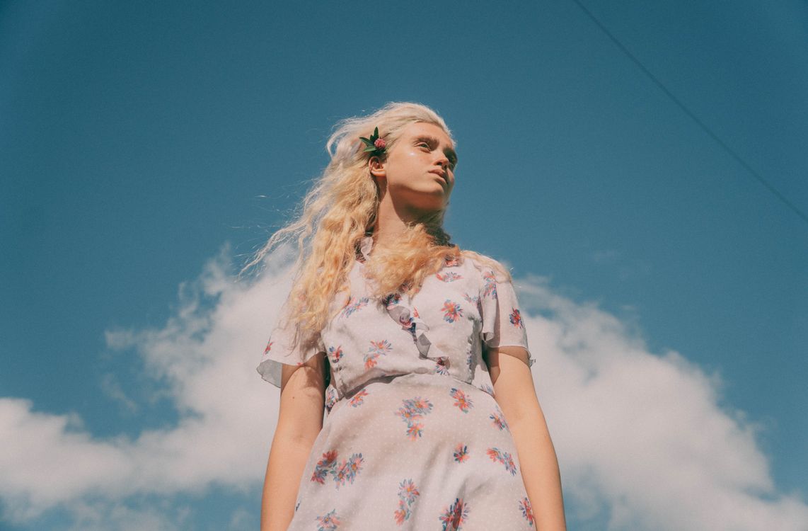Free Low angle of young tender female in dress with floral ornament looking away under cloudy sky Stock Photo