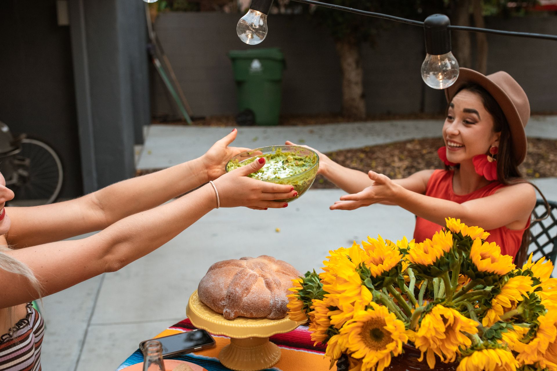 Two women enjoy Mexican cuisine outdoors, exchanging guacamole, surrounded by sunflowers and bread.