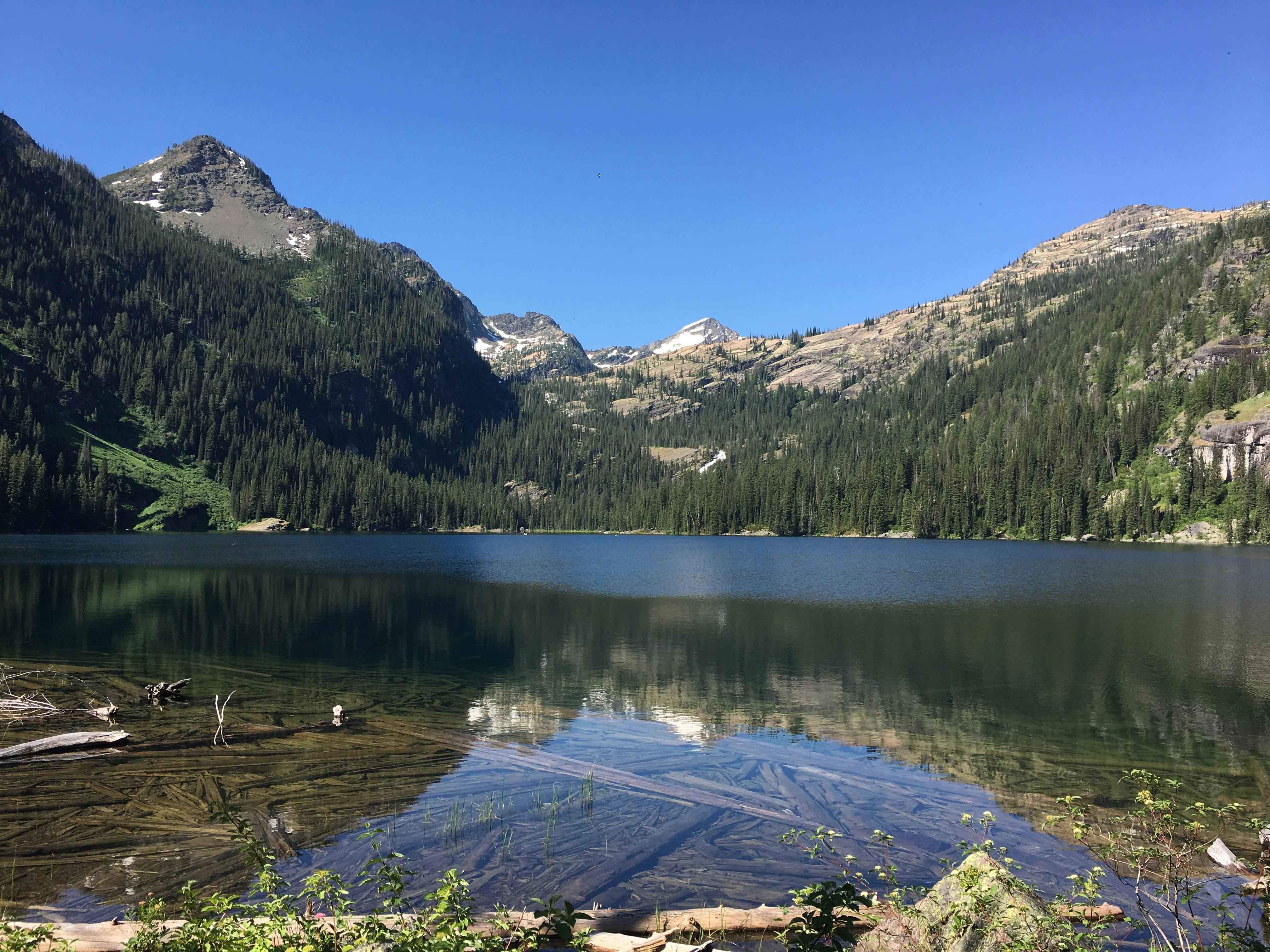 Panoramic view of Seeley Lake with mountain reflections, clear skies, and lush forests.