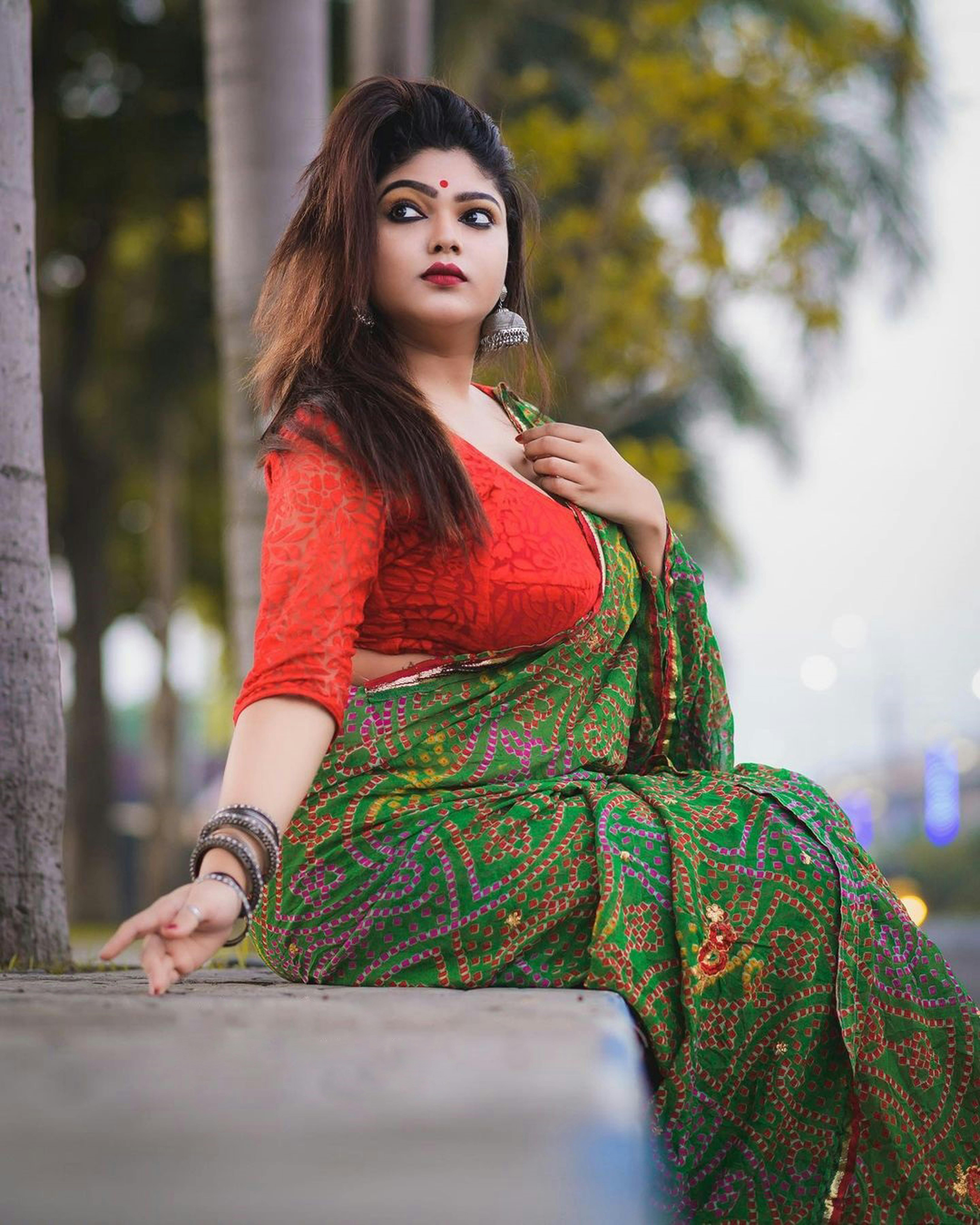 A woman dressed in a colorful saree poses gracefully outdoors, surrounded by nature.