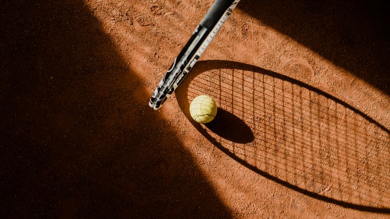 Tennis ball with racket shadow on a sunlit clay court.