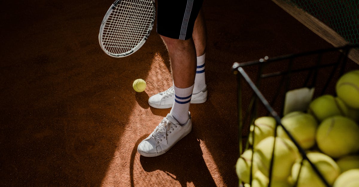 A tennis player stands on a clay court, showing sneakers, racket, and tennis balls in sunlight.