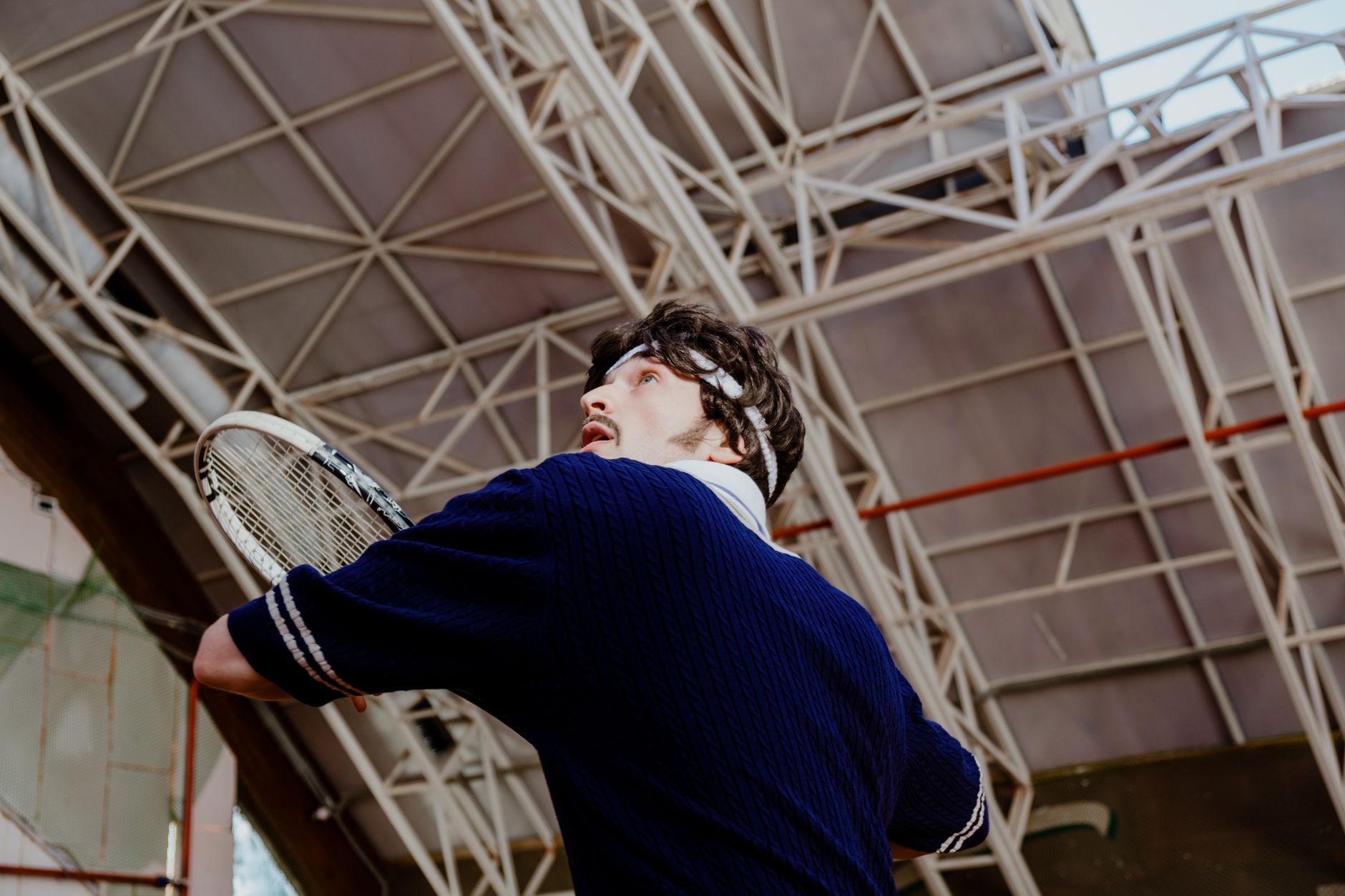 A man in retro attire prepares to hit a tennis ball in an indoor court.