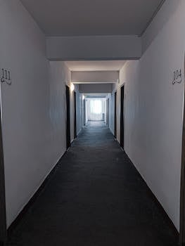 Perspective view of empty hallway with doors and numbers on white walls with window at end of corridor in building