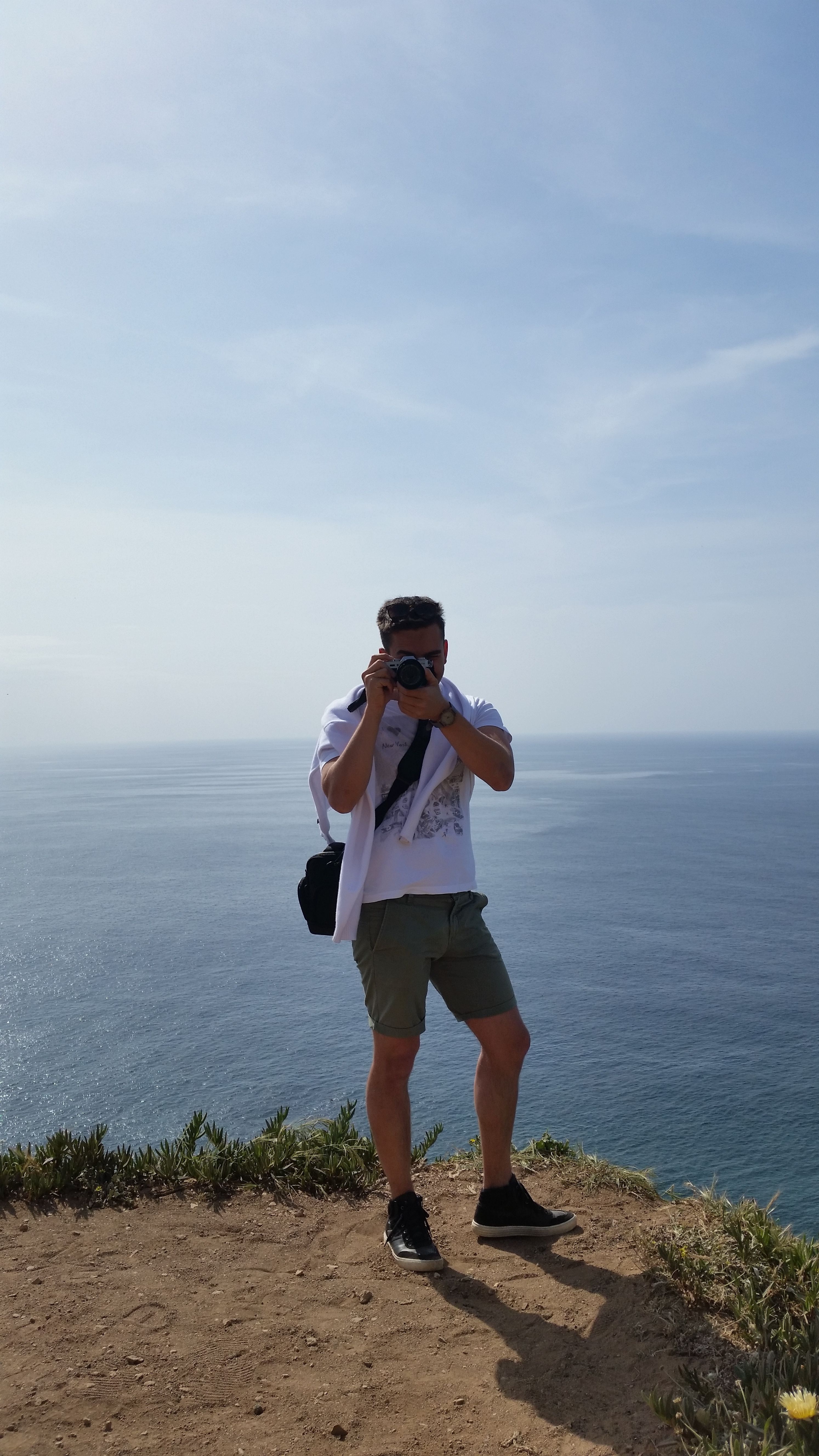 A man stands on a cliff taking photos by the sea in Lisbon.