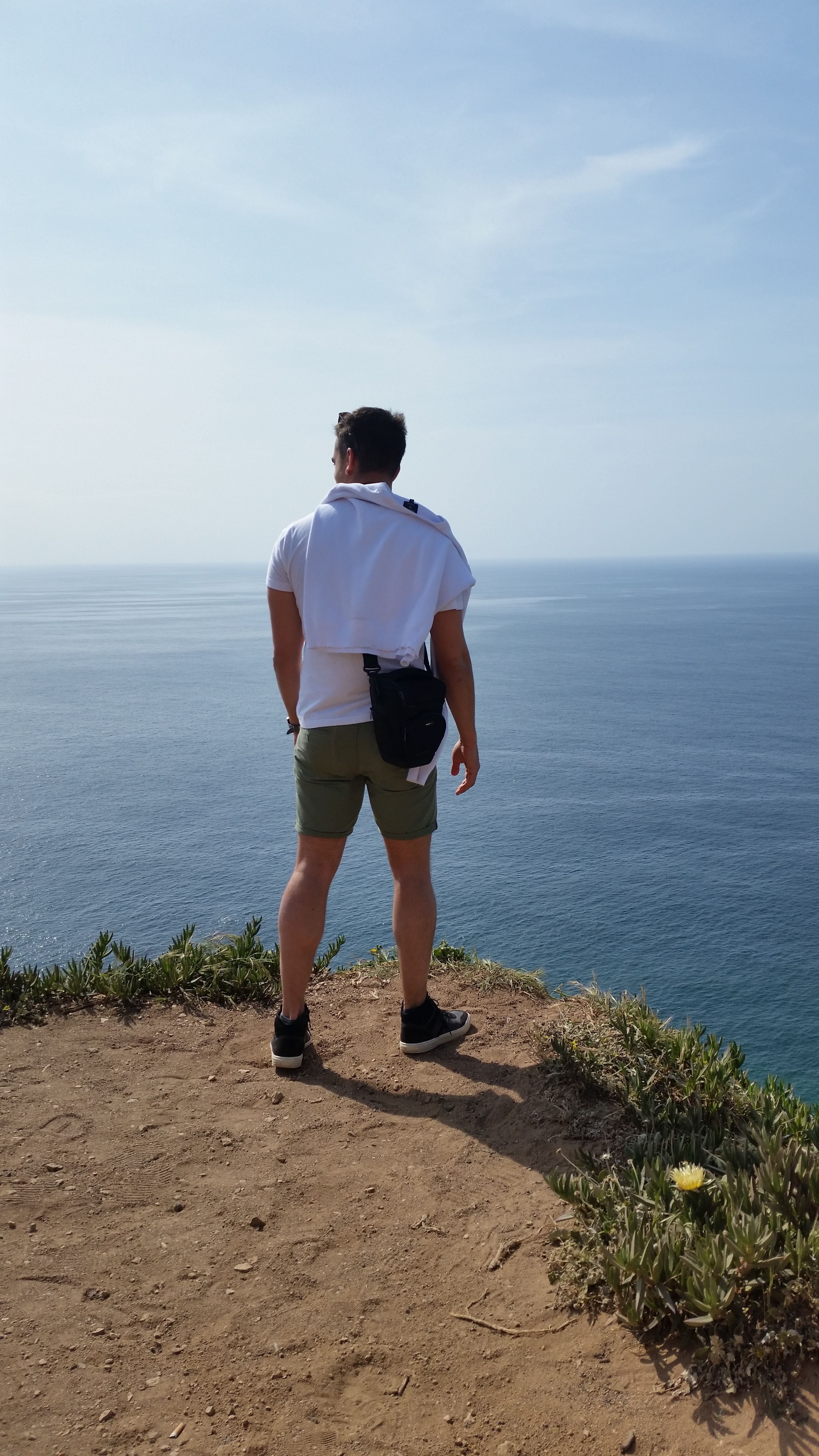 A man standing on a cliff in Lisbon, Portugal, gazing at the Atlantic Ocean on a sunny day.
