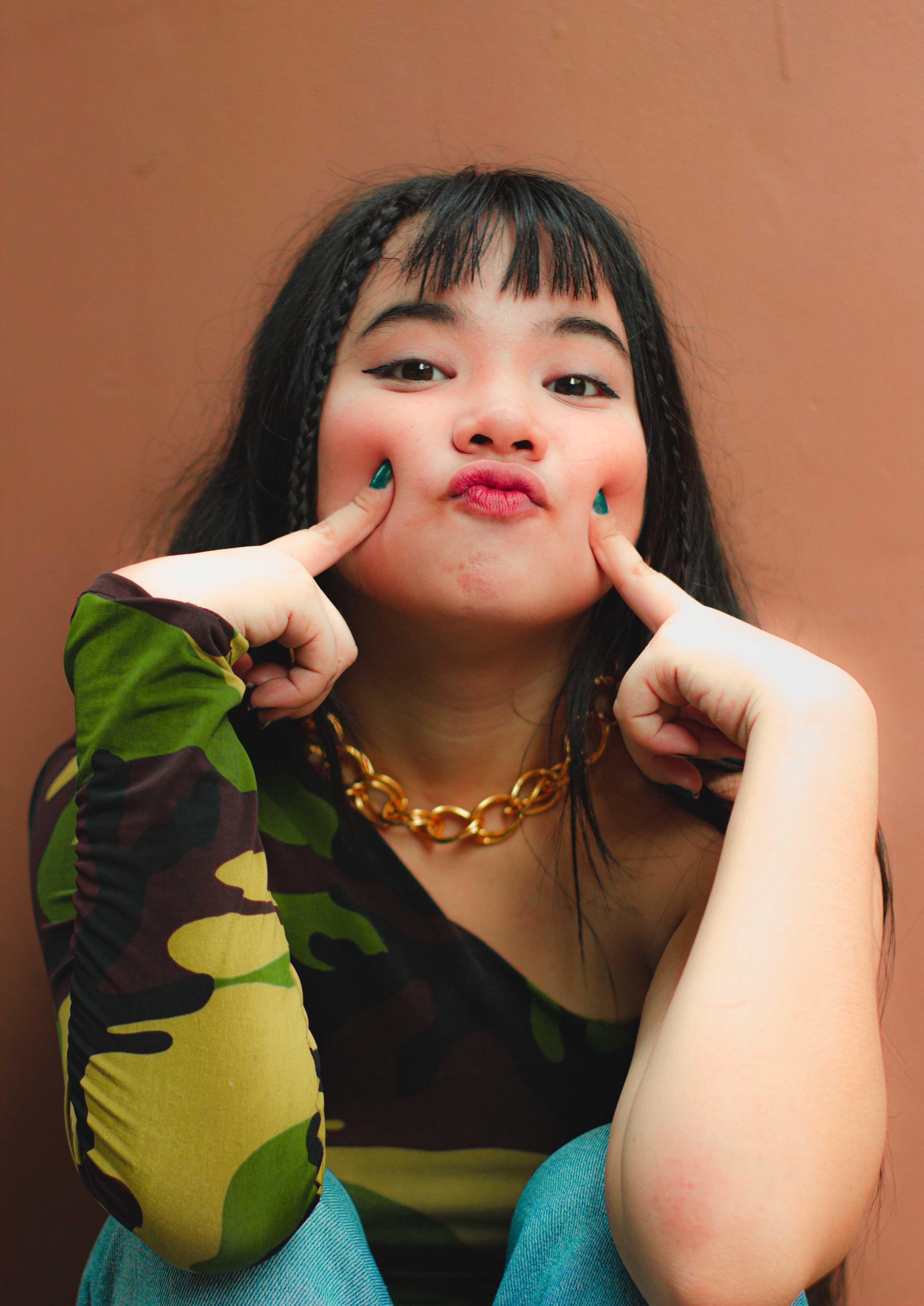 A young woman playfully poses with her fingers on her cheeks, wearing a stylish camouflage shirt.