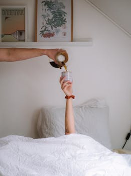 A hand pours coffee using a Chemex over a bed in a minimalist bedroom setting.