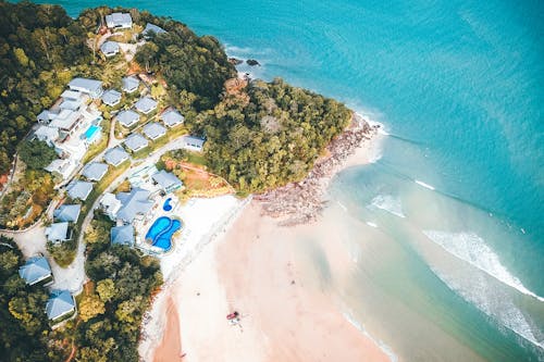 Picturesque aerial view of villas on hill with green tropical trees located on sandy beach of wavy turquoise ocean on sunny day