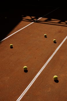 Tennis balls scattered on a sunlit clay court with shadows.