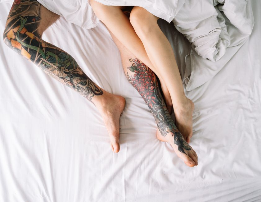 Two tattooed legs intertwined on white bed sheets, creating a cozy and intimate scene.