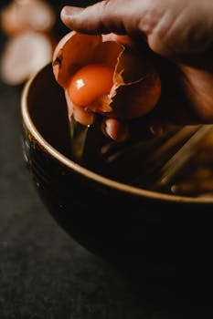 Close-up of a hand cracking a fresh egg into a dark mixing bowl, showcasing the vibrant yolk.